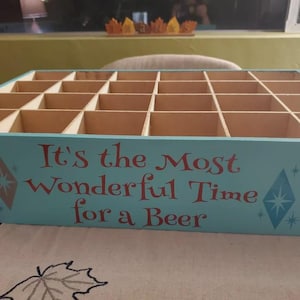 Op de afbeelding: Een lichtblauwe houten adventskalenderdoos met vierentwintig vierkante vakjes. Het voorpaneel bevat rode tekst met de tekst, It's the Most Wonderful Time for a Beer, geflankeerd door twee blauwe en oranje sterontwerpen aan de zijkanten.