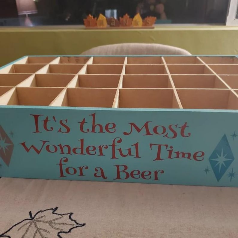 Op de afbeelding: Een lichtblauwe houten adventskalenderdoos met vierentwintig vierkante vakjes. Het voorpaneel bevat rode tekst met de tekst, It's the Most Wonderful Time for a Beer, geflankeerd door twee blauwe en oranje sterontwerpen aan de zijkanten.