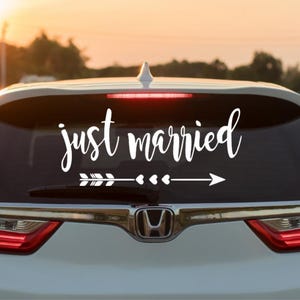 May include: A white car with a "just married" sticker on the back window. The sticker features a white arrow with three hearts and a white arrowhead.