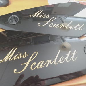 Op de afbeelding: Twee glanzende zwarte, taps toelopende panelen met de tekst Miss Scarlett in een cr&egrave;mekleurig cursief lettertype. Elk paneel is voorzien van een kleine, ronde zwarte knop of sluiting die zich bij het bredere uiteinde van het oppervlak bevindt.