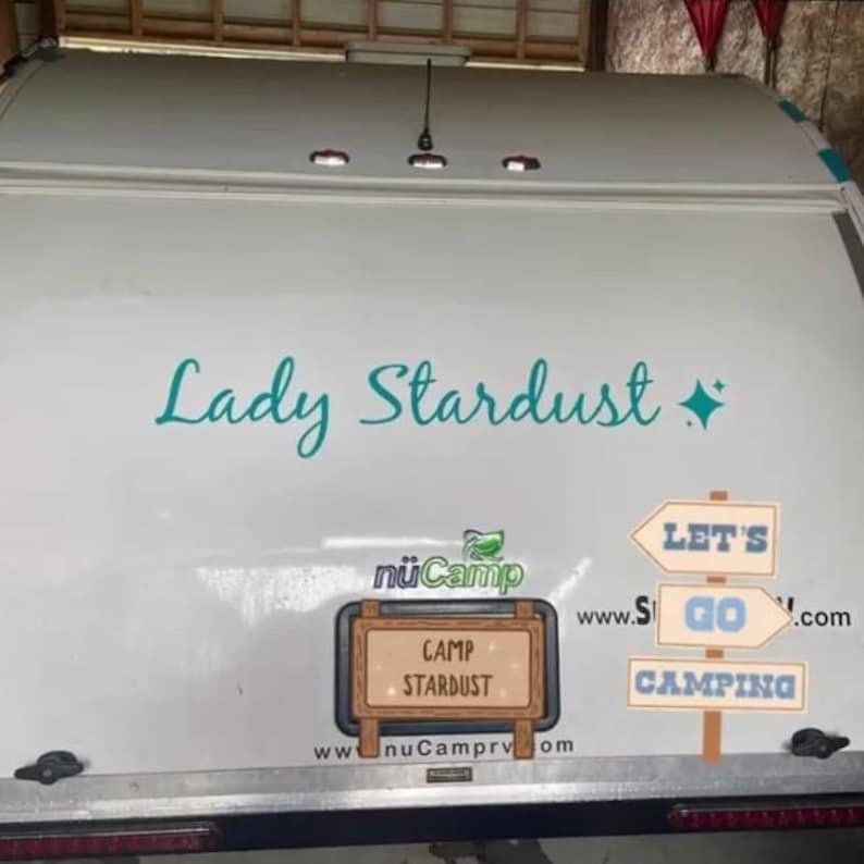 Op de afbeelding: De achterkant van een witte camper met de turquoise tekst Lady Stardust en een sterpictogram. Het bevat een nuCamp-logo, een rechthoekig bord met de tekst Camp Stardust en een bord in drie delen met de blauwe tekst Let's Go Camping.