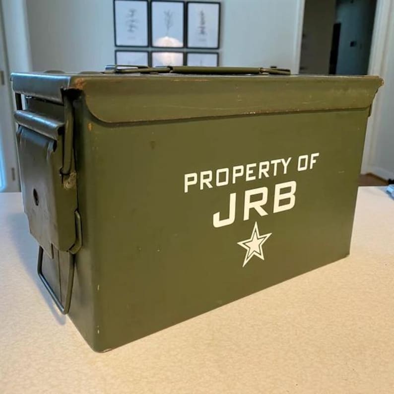 Op de afbeelding: Een olijfgroene metalen munitiekist met een scharnierend deksel, een sluiting en een handvat aan de bovenkant. Op de zijkant staat de witte tekst PROPERTY OF JRB boven een witte afbeelding van een vijfpuntige ster. De kist is rechthoekig.