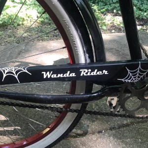 Op de afbeelding: Een zwarte kettingkast voor een fiets met witte spinnenwebafbeeldingen aan beide uiteinden en de tekst Wanda Rider in het wit in het midden gedrukt. De kast bevindt zich boven een zwarte fietsketting, een metalen tandwiel en een wiel met een rode velg.