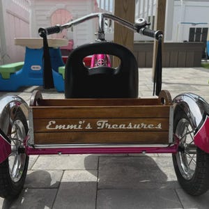 Op de afbeelding: Een roze driewieler met een houten krat aan de achterkant, waarop de tekst Emmi's Treasures in het wit is gedrukt. De driewieler heeft een verchroomd stuur met zwarte handvatten en lange zwarte kwastjes, een zwarte zitting en twee achterwielen.
