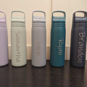 Op de afbeelding: Vijf cilindrische waterflessen met ge&iuml;ntegreerde handgreepdeksels, op een rij geplaatst. De flessen hebben matte afwerkingen in lavendel, lichtgroen, lichtblauw, teal en donkerblauw, elk gepersonaliseerd met verticale namen: Samantha, Jake, Ryan en Brandon.