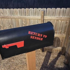 Op de afbeelding: Een zwarte metalen brievenbus gemonteerd op een houten paal, voorzien van een felrode vlag aan de zijkant en de tekst "RETURN TO SENDER" in dikke rode letters op het voorpaneel. De brievenbus heeft een matte afwerking en een rechthoekige vorm.