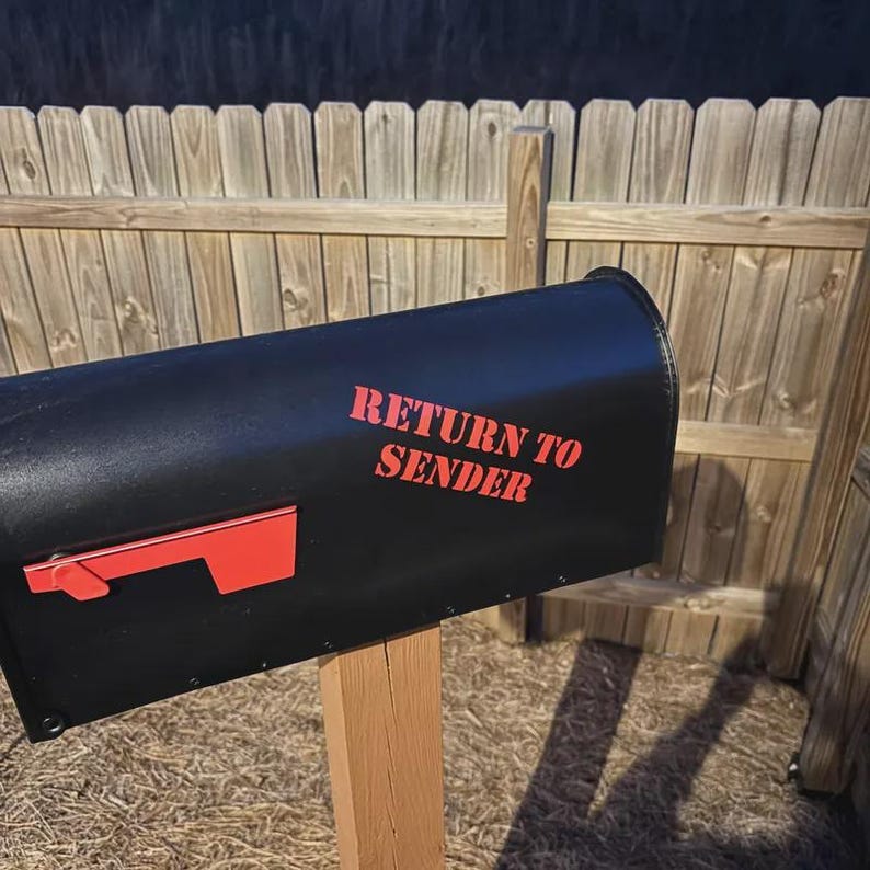 Op de afbeelding: Een zwarte metalen brievenbus gemonteerd op een houten paal, voorzien van een felrode vlag aan de zijkant en de tekst "RETURN TO SENDER" in dikke rode letters op het voorpaneel. De brievenbus heeft een matte afwerking en een rechthoekige vorm.