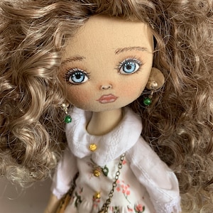 May include: A handmade doll with brown curly hair, blue eyes, and a white dress with floral print. The doll is wearing a brown leather purse and has green earrings.