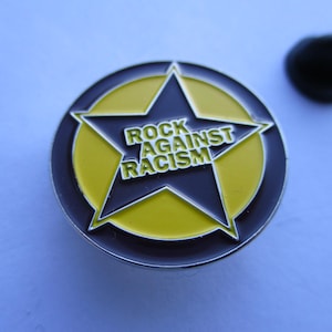 May include: A circular enamel pin featuring a dark brown star set against a yellow background. The text ROCK AGAINST RACISM is printed in white capital letters across the centre of the star. The pin includes a separate black metal clutch backing.