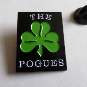 May include: Black and green enamel pin with a shamrock and the text "THE POGUES" in white.