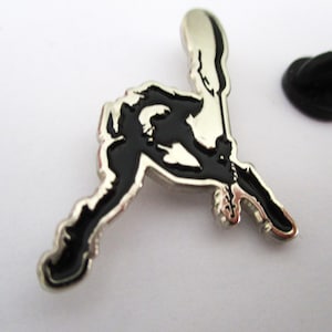 May include: A silver and black metal pin featuring a silhouette of a person playing a guitar. The pin has a safety clasp on the back.