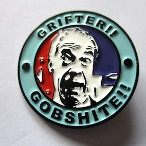 May include: A circular enamel badge with a light blue surround. The badge features a stylized face within a red and blue design, with the words "GRIFTER!!" and "GOBSHITE!!" in black text around the image.