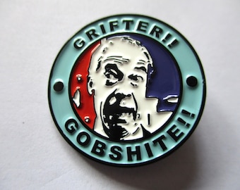 May include: A round enamel pin with a light blue border and a central image of a man's face. The pin features the words "GRIFTER!!" and "GOBSHITE!!" in black text around the image, with red and blue accents.