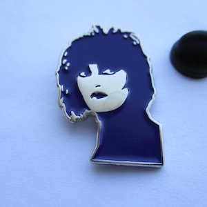 May include: A metal enamel pin featuring a stylised silhouette of a head with dark purple hair and a silver face. The design includes a silver outline and a separate black rubber clutch backing piece positioned to the right of the pin.