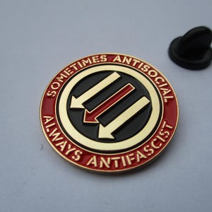 May include: A round metal enamel pin featuring a black centre with three diagonal arrows in gold and red. The outer red border contains the gold text "SOMETIMES ANTISOCIAL ALWAYS ANTIFASCIST". A black rubber clutch backing is positioned to the side.