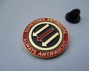 May include: A round metal enamel pin featuring a black center with three diagonal arrows in gold and red. The outer red border contains the gold text "SOMETIMES ANTISOCIAL ALWAYS ANTIFASCIST". A black rubber clutch backing is positioned to the side.