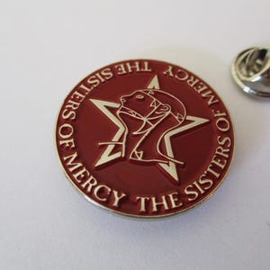 May include: A red and silver pin featuring a five-pointed star and a stylised figure. The text surrounding the star reads "The Sisters of Mercy".