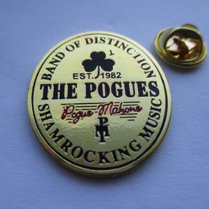 May include: Gold-colored metal pin with a black and white design. The pin features the text "Band of Distinction", "Est. 1982", "The Pogues", "Pogue Mahone", and "Shamrocking Music".