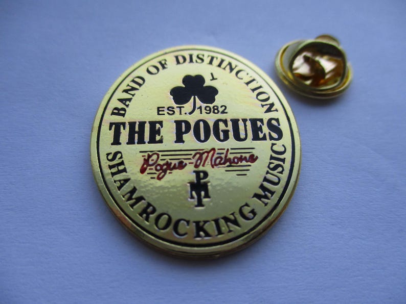 May include: Gold-colored metal pin with a black and white design. The pin features the text "Band of Distinction", "Est. 1982", "The Pogues", "Pogue Mahone", and "Shamrocking Music".