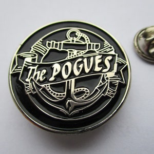 May include: A black and silver metal pin with an anchor and the text "The Pogues" in a banner.