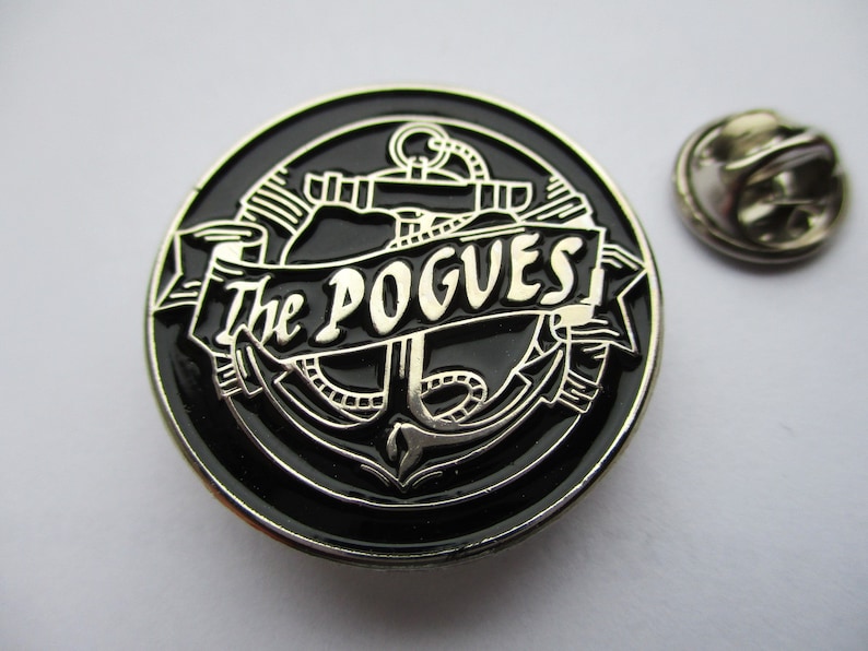 May include: A black and silver metal pin with an anchor and the text "The Pogues" in a banner.