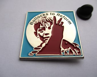 May include: Square enamel pin with a blue border and a silver interior. The pin features a stylized illustration of a person with the text "Bollocks to Reform" in a circular design. A black pin back is next to the pin.