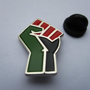 May include: A metal enamel pin shaped as a raised fist, featuring a colour scheme of green, black, and red. The design includes a green forearm, a black wrist, and red fingers. A separate black circular pin back is positioned to the right of the main pin.