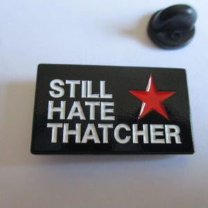 May include: A black enamel pin badge with the words "STILL HATE THATCHER" in white lettering and a red star. A black pin back is also visible. The badge is a political statement.