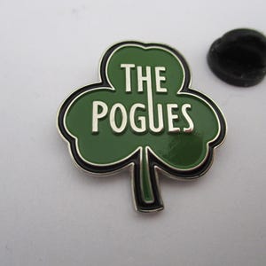 May include: A green shamrock-shaped pin with a silver border and the text "THE POGUES" in white letters.