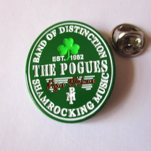 May include: Green and white enamel pin with the text "Band of Distinction" and "The Pogues" in white. The pin also features a green shamrock and the text "Est. 1982" and "Shamrocking Music".