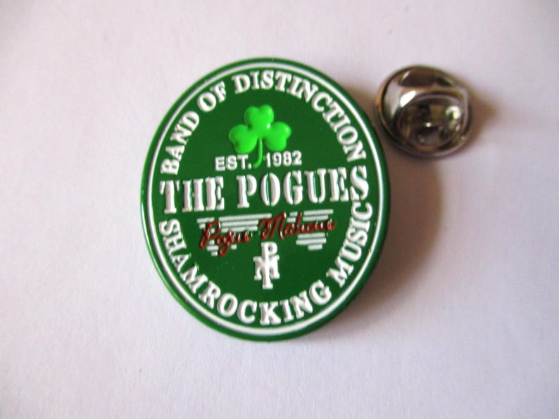 May include: Green and white enamel pin with the text "Band of Distinction" and "The Pogues" in white. The pin also features a green shamrock and the text "Est. 1982" and "Shamrocking Music".