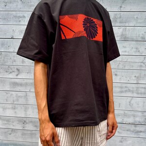 Puede incluir: Una camiseta negra de manga corta con un gráfico rectangular en el pecho. El gráfico muestra un fondo naranja vibrante con finas líneas verticales negras y una gran flor de crisantemo oscura y estilizada en el lado derecho.