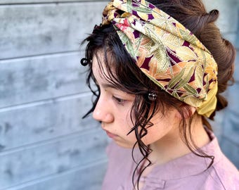Puede incluir: Una diadema de tela anudada con un estampado botánico de hojas verdes, amarillas y naranjas sobre un fondo morado oscuro. La diadema es ancha y fruncida en la parte superior, con una sección de tela amarilla clara lisa visible en la parte posterior.