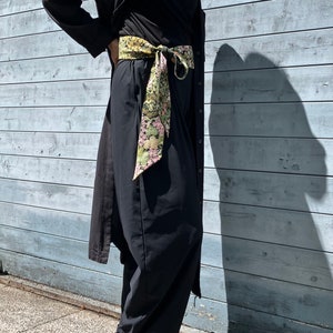 May include: A black long-sleeved shirt with a floral patterned scarf tied around the waist. The shirt has a long, flowing silhouette and is worn with black trousers and white shoes.
