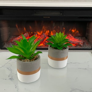 May include: Two potted artificial succulents in grey and white pots with a gold accent. The pots are sitting on a white marble surface in front of a fireplace.