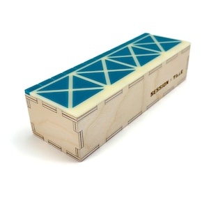 May include: A rectangular wooden box with light-colored plywood sides featuring visible finger joints. The top surface is a smooth, teal-colored acrylic panel with a white grid pattern. The text SESSION x TAAK is printed on the side of the box.