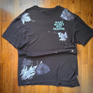 Op de afbeelding: Donkerbruin t-shirt met bladprints in wit en paars. Het shirt heeft de tekst "MAKE MONEY NOT FRIENDS" in turquoise. Het shirt is gemaakt van een zacht materiaal.