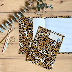 May include: A notebook and cover set with a leopard print design in brown and black. The notebook is open, showing lined paper. The cover has a small label with the text "POP PIP". Pencils and decorative paper clips are also visible on a wooden surface.