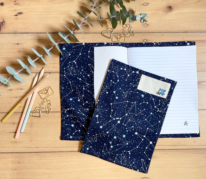 May include: A notebook with a constellation print cover. The notebook is open to reveal lined paper. A small white label with the text "POP CLIP" is attached to the cover.