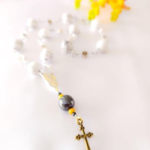 Può includere: Un rosario con perline bianche e una catena d'argento. Il rosario presenta una perlina grigio scuro, perline gialle e un ciondolo a forma di croce color oro. Un fiore giallo è sullo sfondo.