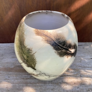 May include: A spherical ceramic vase with a smooth, off-white finish featuring dark, feathery, smoke-like patterns wrapping around the exterior. The vessel has a wide, open circular mouth and a clean, white interior surface visible from the top.