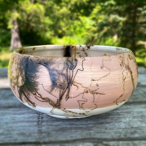 May include: A handcrafted wooden bowl with a unique design. The bowl features a light pink and cream color scheme with intricate black, brown, and yellow patterns. The bowl is round and sits on a wooden surface, showcasing its natural wood grain.