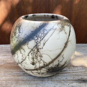 May include: A spherical ceramic vase with a smooth, off-white matte finish. The surface is decorated with intricate, dark black horsehair markings that create thin, chaotic, and linear patterns wrapping around the rounded body of the vessel.