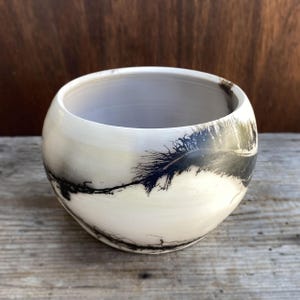 May include: A round, off-white ceramic bowl with a dark, feather-like design. The bowl has a smooth, glossy finish and a wide opening. The interior is a light gray color. The design is a striking contrast against the white background.