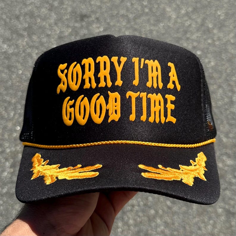 May include: A black trucker hat featuring the yellow embroidered text SORRY I'M A GOOD TIME on the front panel. The hat includes a yellow braided cord across the base of the crown and yellow embroidered leaf accents on the brim.
