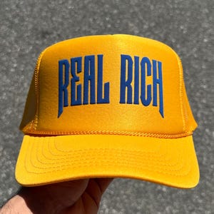 May include: A bright yellow trucker hat featuring the text REAL RICH printed in bold blue capital letters across the front foam panel. The cap includes a structured front, a curved brim with stitching, and a mesh back section for ventilation.
