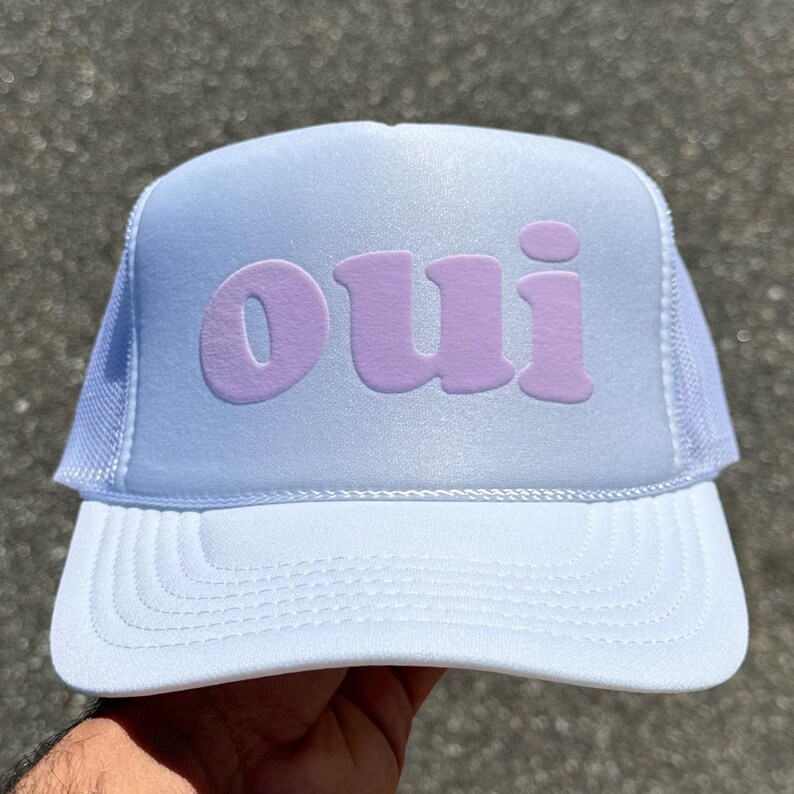 May include: A white trucker hat featuring a foam front panel with the word "oui" printed in a soft, light purple font. The cap includes a structured brim and breathable mesh side and back panels, providing a clean and simple aesthetic design.