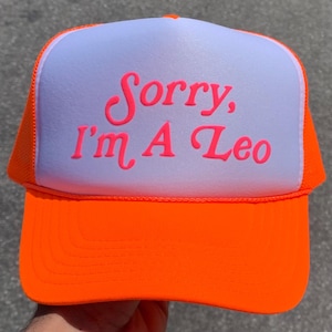 May include: A bright neon orange trucker hat featuring a white front panel. The text "Sorry, I'm A Leo" is printed in a bold, neon pink script font across the center of the white panel. The hat includes a structured brim and mesh side panels.