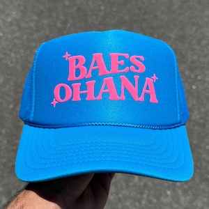 May include: A bright blue trucker hat featuring a foam front panel and mesh back. The front displays the text "BAES OHANA" in bold, hot pink lettering, accented by small four-pointed star symbols positioned above and below the text.