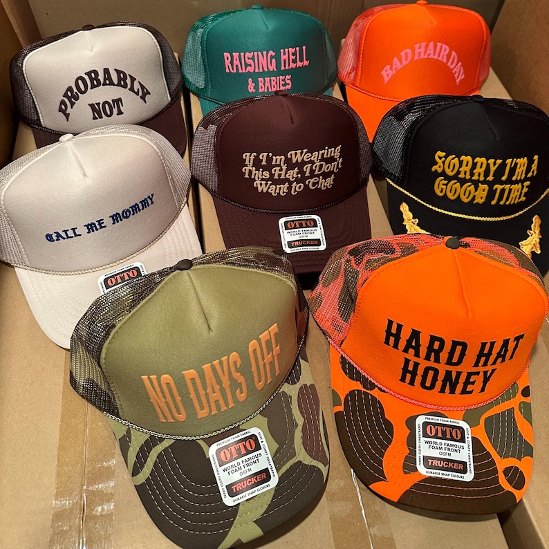 May include: A collection of trucker hats in various colors, including brown, green, orange, and black. Each hat features a different text graphic, such as "Probably Not," "Raising Hell & Babies," and "Hard Hat Honey." The hats have mesh backs and snap closures.